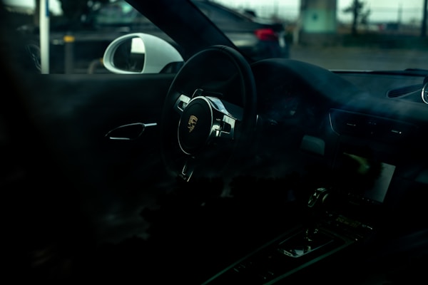 A dark interior of a car with a focus on the steering wheel featuring a prominent car logo. The dashboard and the side mirror are partially visible, and the car's exterior environment can be seen through the windows.