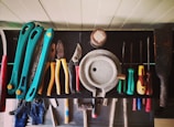 A collection of various hand tools neatly arranged on a black surface. The tools include pliers, screwdrivers, cutters, and a saw with handles in different colors such as turquoise, red, yellow, and green. A round metallic object is centrally placed, possibly a weight or industrial component. The tools appear well-used, hinting at a workshop environment. Below the surface, additional tools and a pair of blue gloves can be observed.