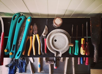 A collection of various hand tools neatly arranged on a black surface. The tools include pliers, screwdrivers, cutters, and a saw with handles in different colors such as turquoise, red, yellow, and green. A round metallic object is centrally placed, possibly a weight or industrial component. The tools appear well-used, hinting at a workshop environment. Below the surface, additional tools and a pair of blue gloves can be observed.