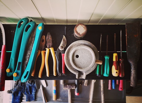 A collection of various hand tools neatly arranged on a black surface. The tools include pliers, screwdrivers, cutters, and a saw with handles in different colors such as turquoise, red, yellow, and green. A round metallic object is centrally placed, possibly a weight or industrial component. The tools appear well-used, hinting at a workshop environment. Below the surface, additional tools and a pair of blue gloves can be observed.