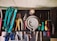 A collection of various hand tools neatly arranged on a black surface. The tools include pliers, screwdrivers, cutters, and a saw with handles in different colors such as turquoise, red, yellow, and green. A round metallic object is centrally placed, possibly a weight or industrial component. The tools appear well-used, hinting at a workshop environment. Below the surface, additional tools and a pair of blue gloves can be observed.