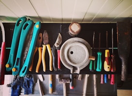 A collection of various hand tools neatly arranged on a black surface. The tools include pliers, screwdrivers, cutters, and a saw with handles in different colors such as turquoise, red, yellow, and green. A round metallic object is centrally placed, possibly a weight or industrial component. The tools appear well-used, hinting at a workshop environment. Below the surface, additional tools and a pair of blue gloves can be observed.