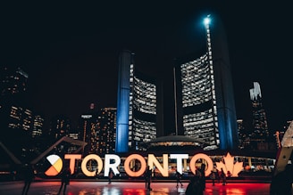 A nighttime cityscape highlighted by illuminated letters spelling out 'Toronto' in front of a modern building, with a mix of people walking around. The background features tall, brightly lit skyscrapers, contributing to the lively urban atmosphere.