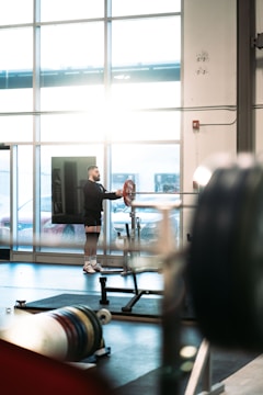 A man stands near a large window in a gym, handling a barbell with weights on a squat rack. The room is bathed in natural light, with gym equipment scattered around. The atmosphere appears calm and focused.