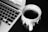 A black and white image features a ceramic cup filled with a hot beverage placed on a dark surface next to a laptop keyboard. The cup has a textured design with a prominent handle, and slight steam is rising from the drink.