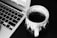 A black and white image features a ceramic cup filled with a hot beverage placed on a dark surface next to a laptop keyboard. The cup has a textured design with a prominent handle, and slight steam is rising from the drink.