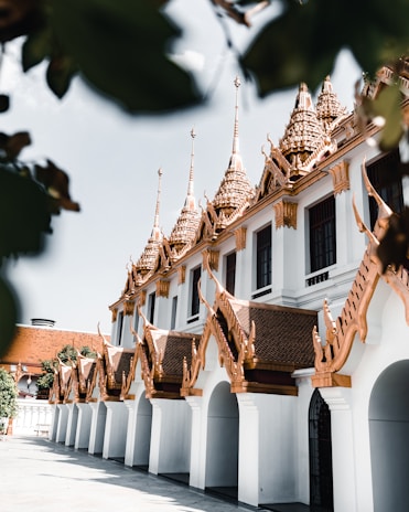 A traditional Asian building featuring intricate, golden spires and ornate architecture with white arches. The design includes detailed patterns, and the bright sunlight enhances the contrast between the gold and white elements. Leaves partially frame the scene in the top corners.