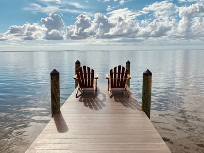 two Adirondack chairs at end of brown wooden dock on calm water under blue sky during daytime