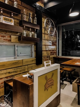 The image depicts a cozy interior with wooden walls, shelves displaying empty glass bottles, and a rustic sign reading 'SELF SERVICE.' A bicycle wheel is mounted on the wall as decoration. A countertop below has a sign that reads 'Pay First, SELF SERVICE,' and a logo featuring a burger is displayed. The setting includes stools and tables, suggesting a small eatery or cafe.