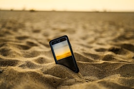 A smartphone is placed upright in the sand of a desert, displaying an image of a sunset with a yellow and orange sky. The smooth, wavy sand surface surrounds the phone. The background features a blurred horizon, evoking a sense of vastness.