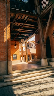 A sunlit architectural space featuring warm brick walls and wooden beams. The scene includes several black hanging lanterns and an arrangement of steps leading up to an open area. The shadows cast intricate patterns on the stone pavement, and there's a glimpse of rooftops in the background.