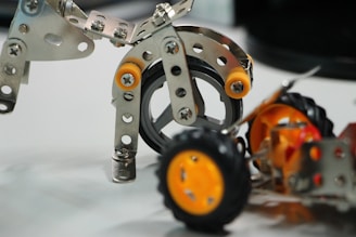 Metal construction parts assembled into a small robotic figure, featuring wheels and bolts. The metal pieces are shiny and perforated, and the assembly includes bright orange elements providing color contrast.