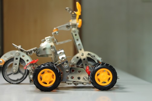 A small model vehicle constructed with metallic parts and orange wheels is placed on a white surface. The structure includes riveted metal beams and plastic gears, appearing like an assembly from a construction toy set. The background is out of focus, highlighting the model.