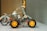 A small model vehicle constructed with metallic parts and orange wheels is placed on a white surface. The structure includes riveted metal beams and plastic gears, appearing like an assembly from a construction toy set. The background is out of focus, highlighting the model.
