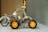 A small model vehicle constructed with metallic parts and orange wheels is placed on a white surface. The structure includes riveted metal beams and plastic gears, appearing like an assembly from a construction toy set. The background is out of focus, highlighting the model.