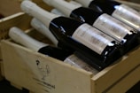 Several bottles of wine with white labels are neatly arranged inside a wooden crate. The crate features an illustration of a rooster and some branding text. The bottles are dark in color with bright labels.