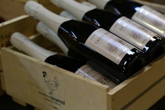 Several bottles of wine with white labels are neatly arranged inside a wooden crate. The crate features an illustration of a rooster and some branding text. The bottles are dark in color with bright labels.