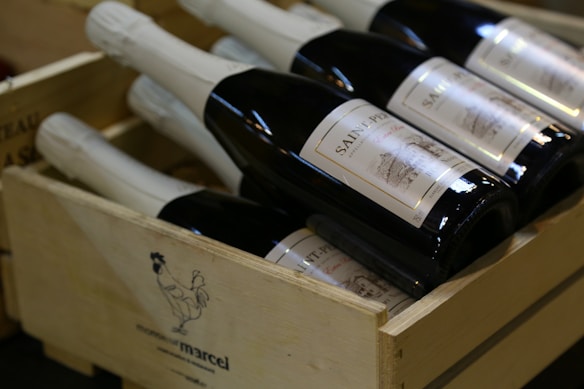 Several bottles of wine with white labels are neatly arranged inside a wooden crate. The crate features an illustration of a rooster and some branding text. The bottles are dark in color with bright labels.