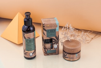 A display of skincare products with CBD ingredients in dark containers and eco-friendly packaging. These include a pump bottle, a small jar, and a box, arranged neatly on a light surface. Nearby is a decorative dried plant and a yellow geometric shape, likely made of paper or cardboard.