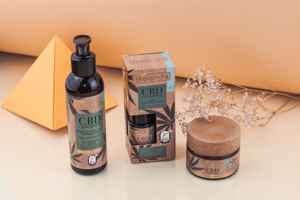 A display of skincare products with CBD ingredients in dark containers and eco-friendly packaging. These include a pump bottle, a small jar, and a box, arranged neatly on a light surface. Nearby is a decorative dried plant and a yellow geometric shape, likely made of paper or cardboard.