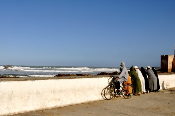 A group of diverse people riding electric and conventional bikes along the sunny coastline of Costa da Caparica, with the ocean in the background.