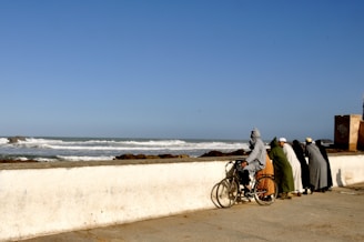 A group of diverse people riding electric and conventional bikes along the sunny coastline of Costa da Caparica, with the ocean in the background.