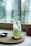 A tall glass filled with a light green beverage sits on a round wooden coaster atop a larger wooden board. The drink is garnished with a sprig of rosemary, and a spoon is placed beside the glass on the board. The background features a blurred window, allowing natural light to softly illuminate the scene.