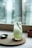 A tall glass filled with a light green beverage sits on a round wooden coaster atop a larger wooden board. The drink is garnished with a sprig of rosemary, and a spoon is placed beside the glass on the board. The background features a blurred window, allowing natural light to softly illuminate the scene.