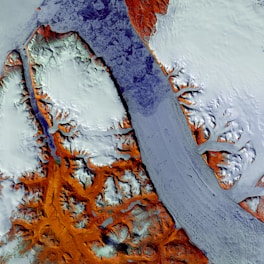 A satellite view of a landscape featuring rugged, snow-covered terrain with contrasting elements of exposed earth in shades of rust and orange. A river or glacier-like formation moves across the frame, winding through the natural formations.