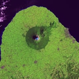A satellite image of a mountain surrounded by lush green vegetation. The mountain features a clearly defined crater and is situated near a coastline with dark blue waters. The topography of the landscape shows various textures, indicating different land uses or types of vegetation.