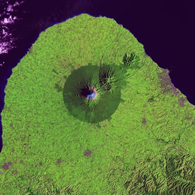 A satellite image of a mountain surrounded by lush green vegetation. The mountain features a clearly defined crater and is situated near a coastline with dark blue waters. The topography of the landscape shows various textures, indicating different land uses or types of vegetation.