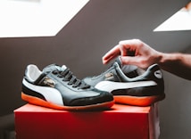 A pair of black and white sneakers with orange soles is placed on a red box. A hand is adjusting one of the shoes, suggesting they might be newly purchased or being put away.