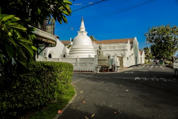 A peaceful street scene features a pristine white stupa as the focal point, complemented by an intricately designed wall with arched patterns. Surrounding greenery and trees add a touch of nature while a clear blue sky enhances the serene atmosphere. A statue of a seated figure, possibly Buddha, is placed near the entrance to the religious site. A few vehicles and pedestrians are visible on the road adjacent to the temple complex.