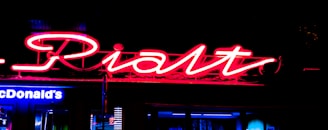 Bright neon signs dominate the dark background, with a red handwritten-style sign overlaying a partially visible blue McDonald's sign below.