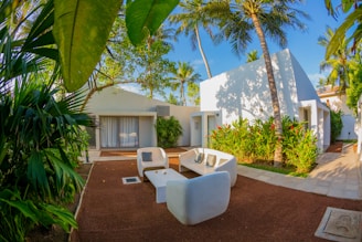 A modern outdoor seating area surrounded by lush tropical vegetation and white, minimalistic buildings. The scene is framed by large palm leaves and features contemporary white furniture arranged on a reddish-brown patio. Tall palm trees and flowering plants enhance the tropical ambiance.