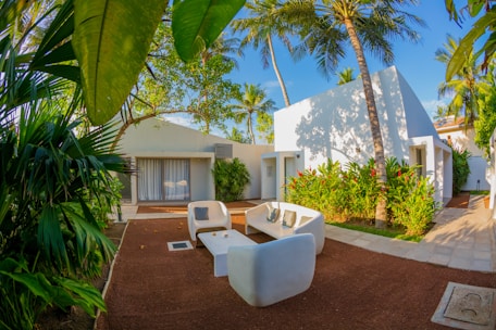 A modern outdoor seating area surrounded by lush tropical vegetation and white, minimalistic buildings. The scene is framed by large palm leaves and features contemporary white furniture arranged on a reddish-brown patio. Tall palm trees and flowering plants enhance the tropical ambiance.