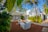 A modern outdoor seating area surrounded by lush tropical vegetation and white, minimalistic buildings. The scene is framed by large palm leaves and features contemporary white furniture arranged on a reddish-brown patio. Tall palm trees and flowering plants enhance the tropical ambiance.