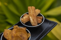 Deep-fried slices of yucca and plantain chips arranged in small black bowls. The background consists of a blurred green, leafy texture, highlighting the golden brown colors of the fried snacks.