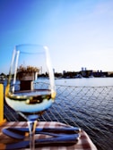 A glass of white wine is positioned in the foreground on a checkered tablecloth, with silverware visible nearby. In the background, there is a waterfront scene featuring a calm body of water, boats, and trees. A clear blue sky dominates the upper half of the image, contributing to a serene outdoor dining atmosphere.