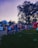A group of people gathered outdoors in what appears to be a fair or festival setting, with green grass and a large tree casting shadows in the background. Various stalls and a small amusement ride are visible, and people are casually mingling, dressed in summer attire.