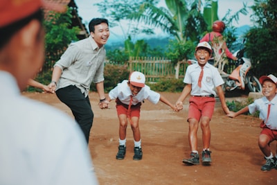 A group of children wearing school uniforms are holding hands and engaging in an outdoor activity with an adult. They appear to be happy and are smiling, with lush greenery and a rural setting in the background.