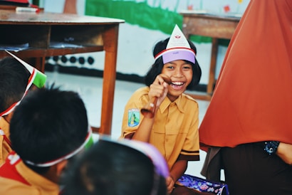 A group of children is gathered in a classroom setting, with one child in focus smiling broadly and holding a wooden object. The children are wearing uniforms and colorful headbands. A wooden table is visible in the background, along with some classroom furniture.