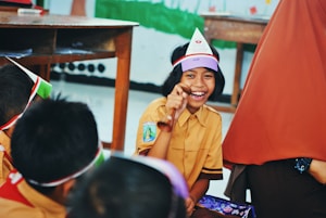 A group of children is gathered in a classroom setting, with one child in focus smiling broadly and holding a wooden object. The children are wearing uniforms and colorful headbands. A wooden table is visible in the background, along with some classroom furniture.