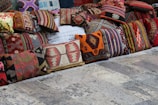 A collection of colorful, intricately patterned cushions displayed on a rustic stone floor. The cushions are arranged in a row and feature various traditional and geometric designs in shades of red, orange, green, brown, and beige.