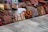 A collection of colorful, intricately patterned cushions displayed on a rustic stone floor. The cushions are arranged in a row and feature various traditional and geometric designs in shades of red, orange, green, brown, and beige.