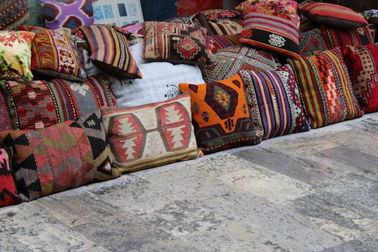 A collection of colorful, intricately patterned cushions displayed on a rustic stone floor. The cushions are arranged in a row and feature various traditional and geometric designs in shades of red, orange, green, brown, and beige.
