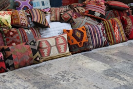 A collection of colorful, intricately patterned cushions displayed on a rustic stone floor. The cushions are arranged in a row and feature various traditional and geometric designs in shades of red, orange, green, brown, and beige.