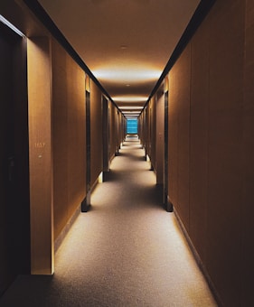 A long, dimly lit corridor with warm lighting that creates a symmetric and inviting atmosphere. The walls are lined with doors, and there is a subtle, modern aesthetic with clean lines and muted colors. At the end of the hallway, a blue glass window provides a focal point.