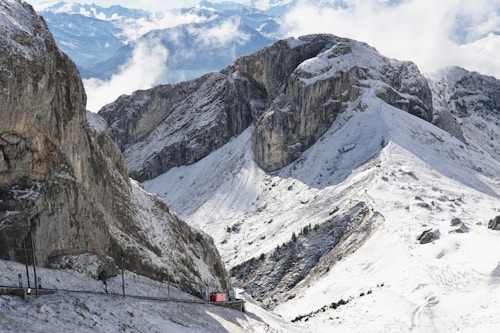 A snowy mountain landscape with rugged peaks and a narrow red train passing through the scene. The mountains are covered with snow, and clouds partially obscure the sky, adding depth to the scenery.