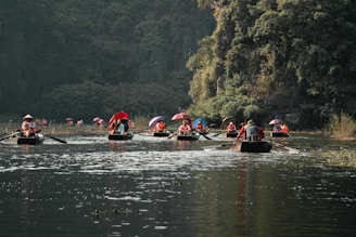 Several small boats filled with people wearing life jackets are rowing on a tranquil river with lush greenery on the banks. Some passengers are holding colorful umbrellas. The scenery is framed by dense forest and rocky cliffs, creating a serene and picturesque setting.